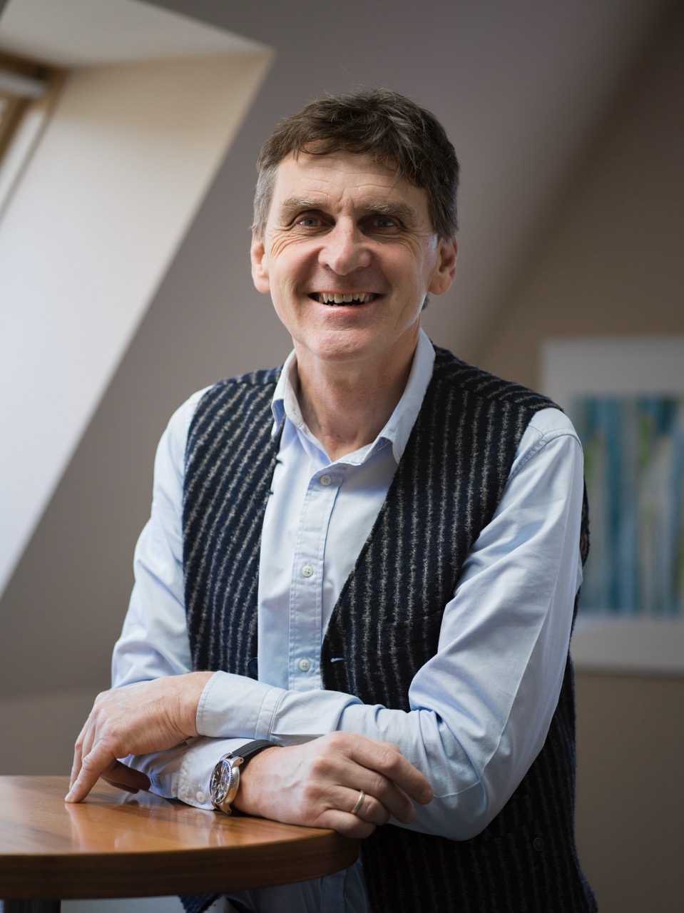 Smiling middle-aged man in light blue shirt and striped vest sitting at a wooden table in a bright room.