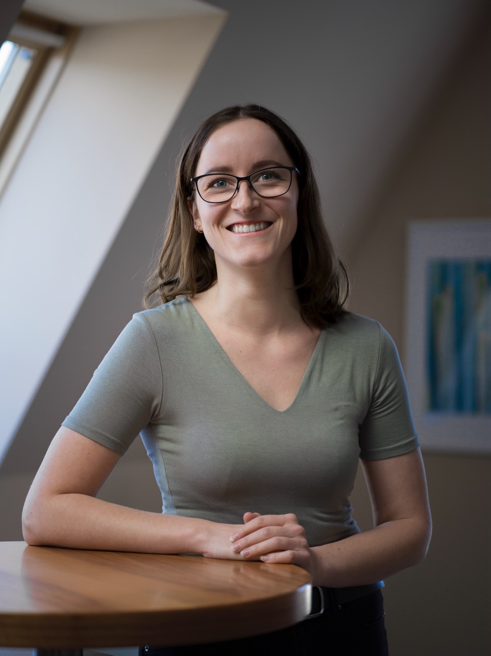 Smiling woman with glasses and shoulder-length brown hair leaning on a wooden table indoors.