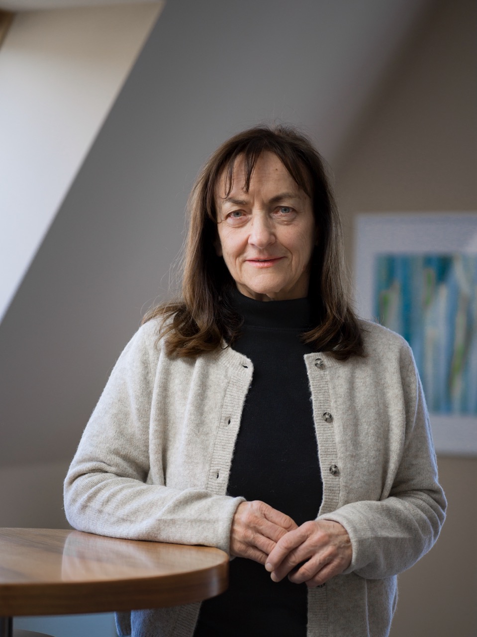 Middle-aged woman with shoulder-length brown hair wearing a beige cardigan over a black top, standing indoors near a wooden table.