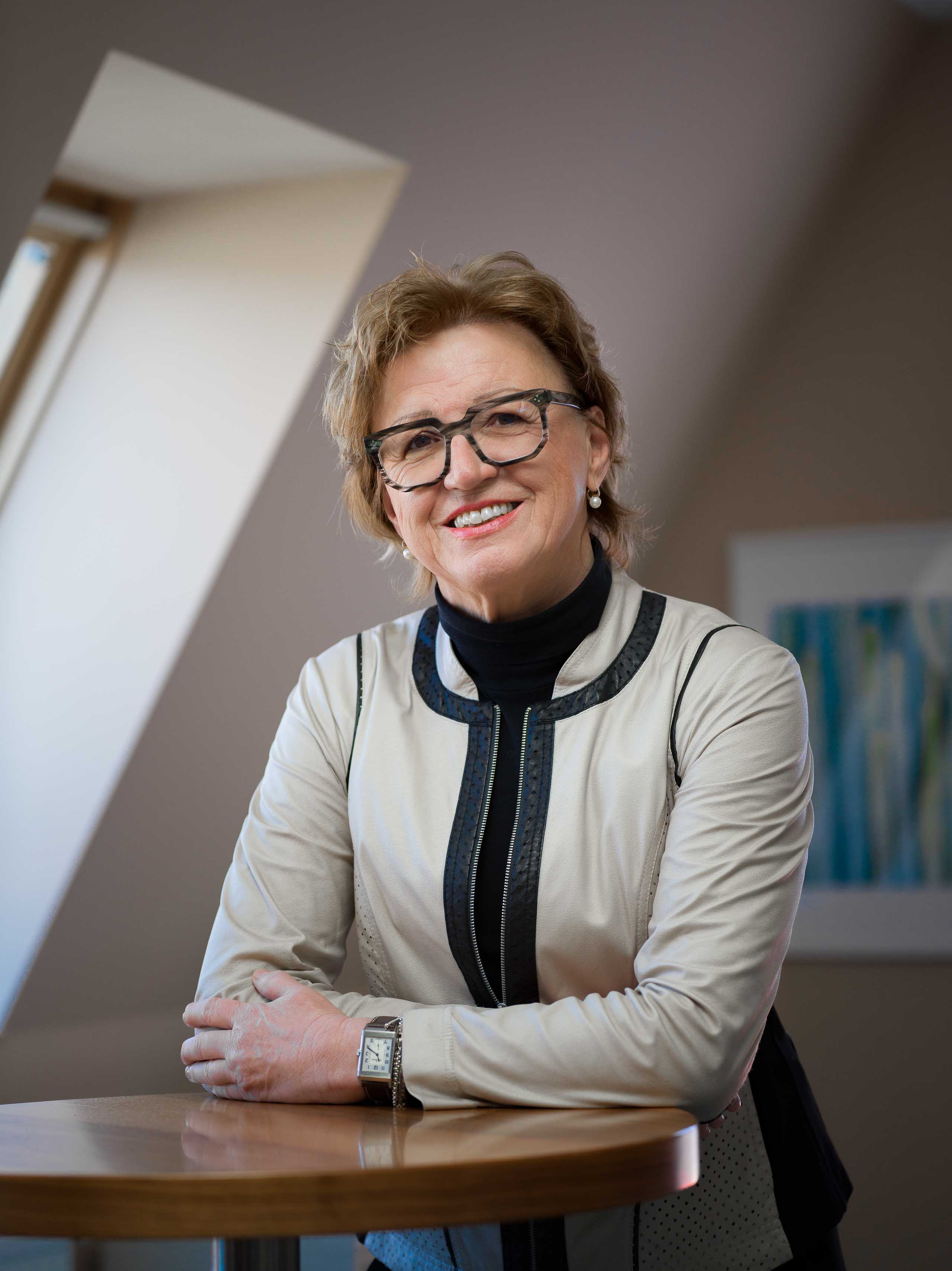 Smiling older woman with glasses and short hair, wearing a beige jacket with black accents, sitting at a wooden table.