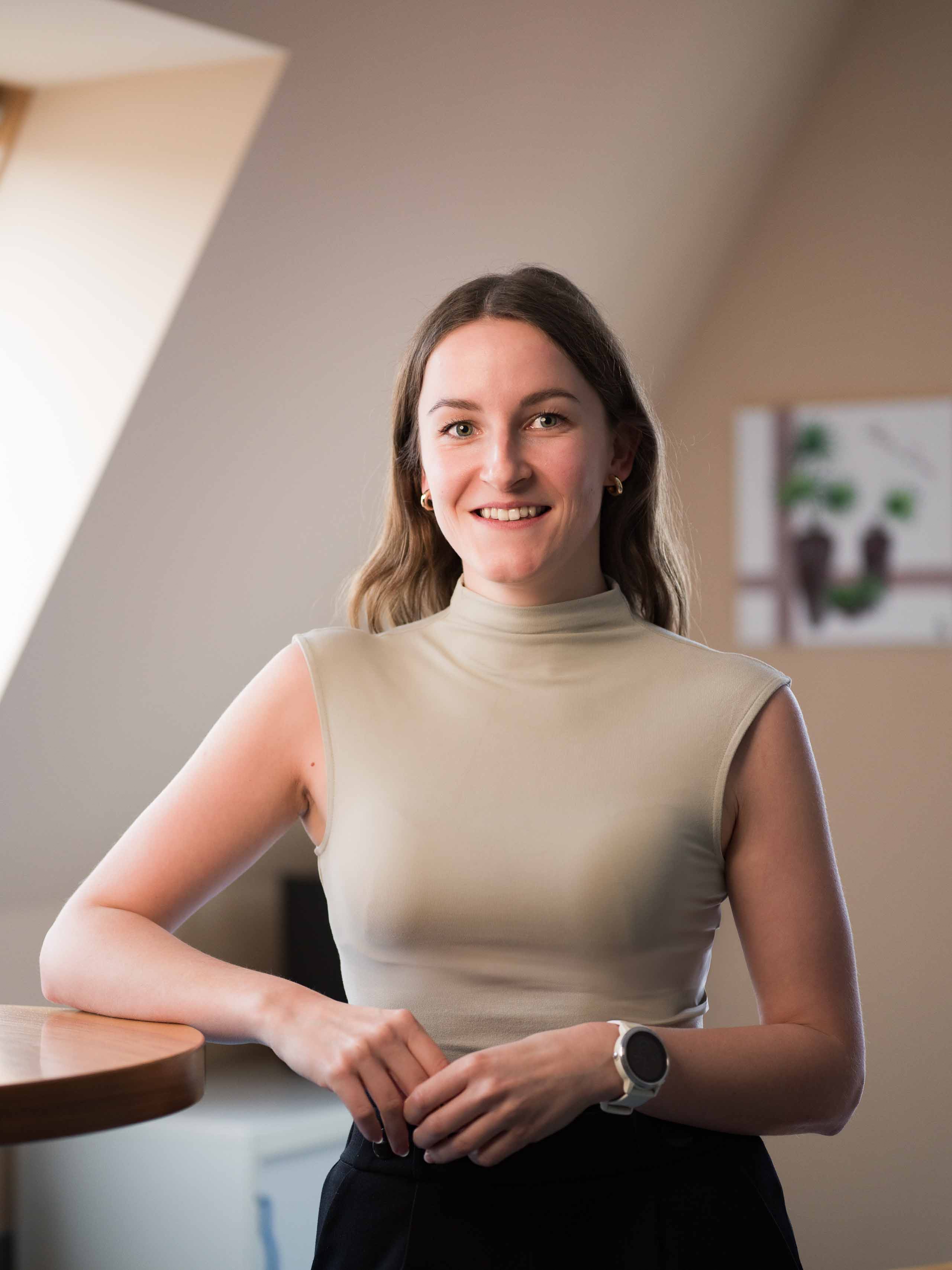 Smiling woman with long hair in a sleeveless beige top and smartwatch, standing indoors near a wooden table.