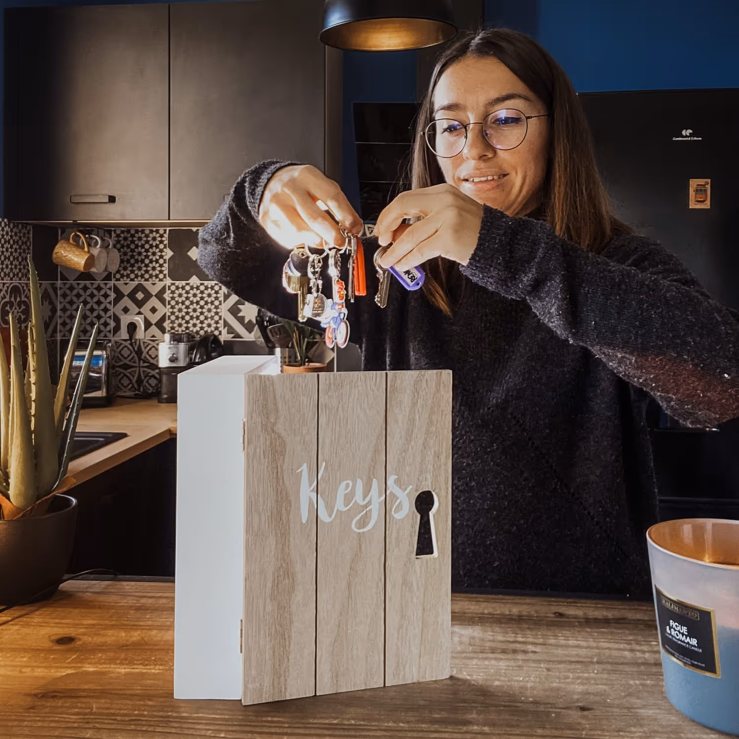 Femme souriante rangeant un trousseau de clés dans une boîte en bois étiquetée 'Keys' sur une table en bois dans une cuisine moderne.