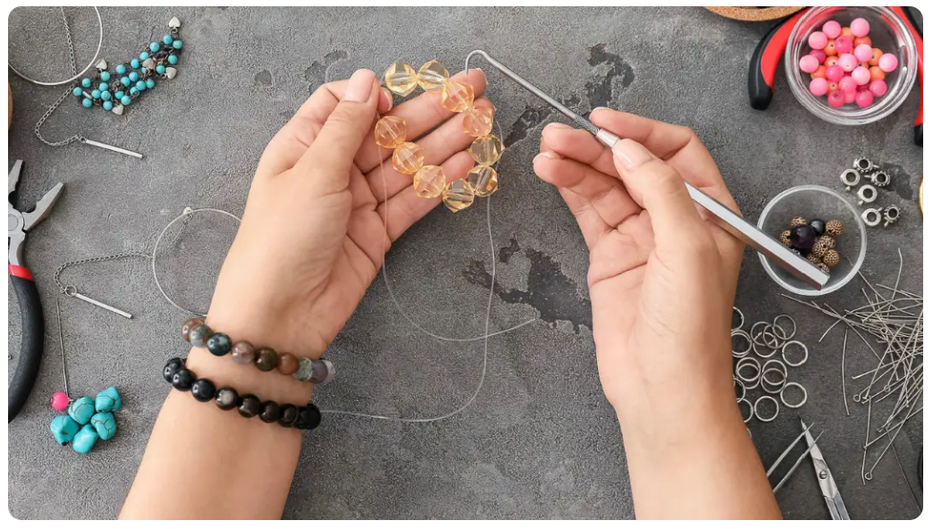 A gray workspace has scattered jewelry supplies. Two hands in the middle are making a bracelet with beads.