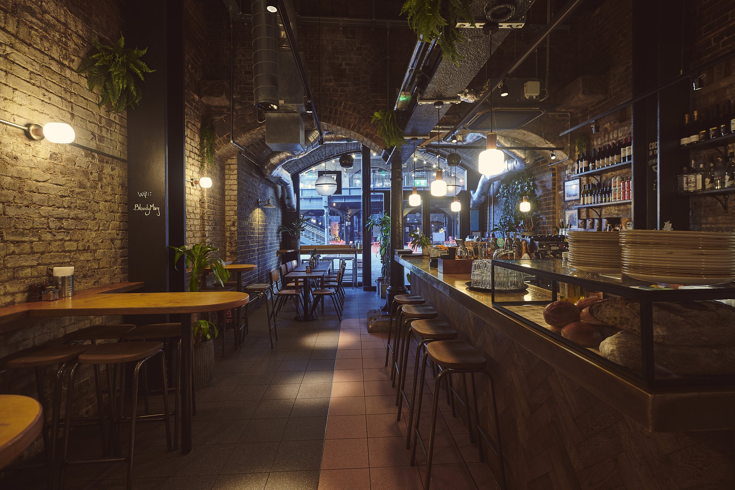 Cozy bar and restaurant interior with wooden stools, tables, brick walls, plants, and hanging lights.