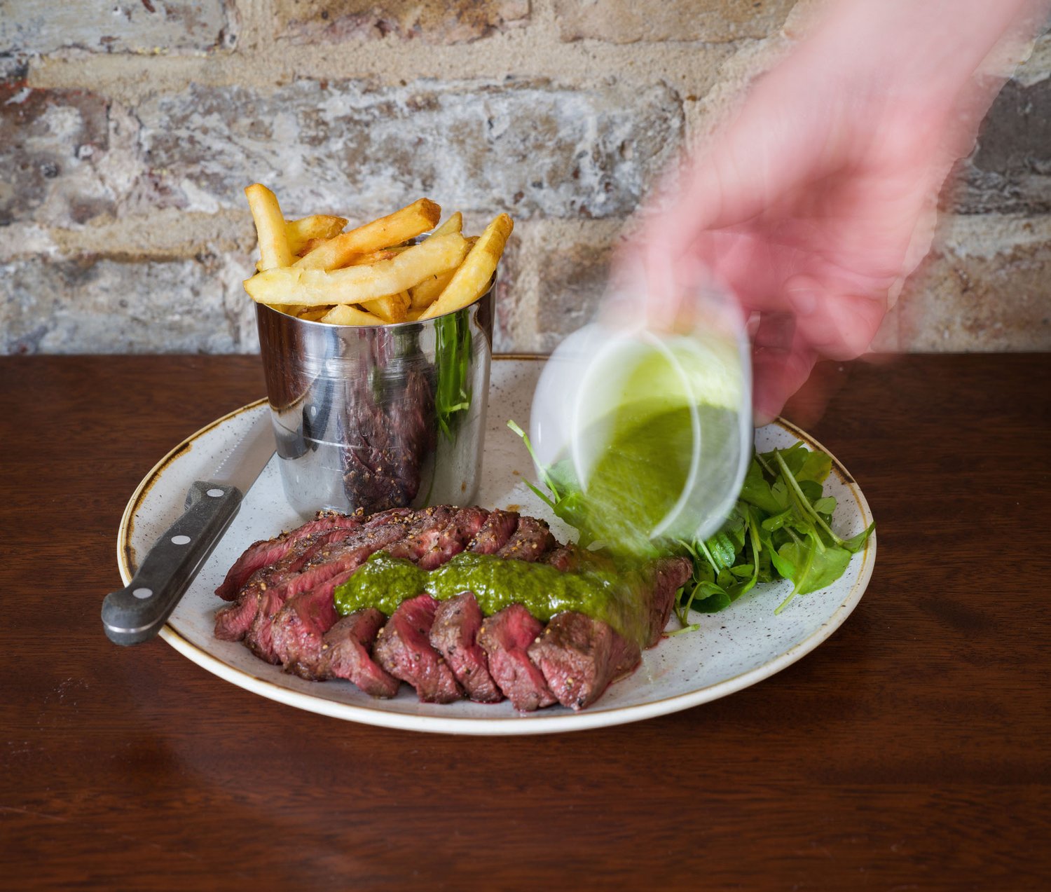 Sliced steak with green sauce being poured, fries, and green salad on plate.