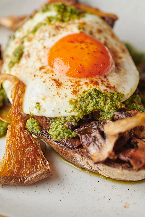 Fried egg with runny yolk on toast topped with mushrooms and green herb sauce on a plate.