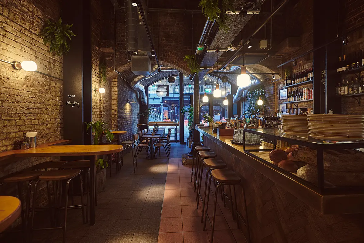 Cozy bar and restaurant interior with wooden stools, tables, brick walls, plants, and hanging lights.