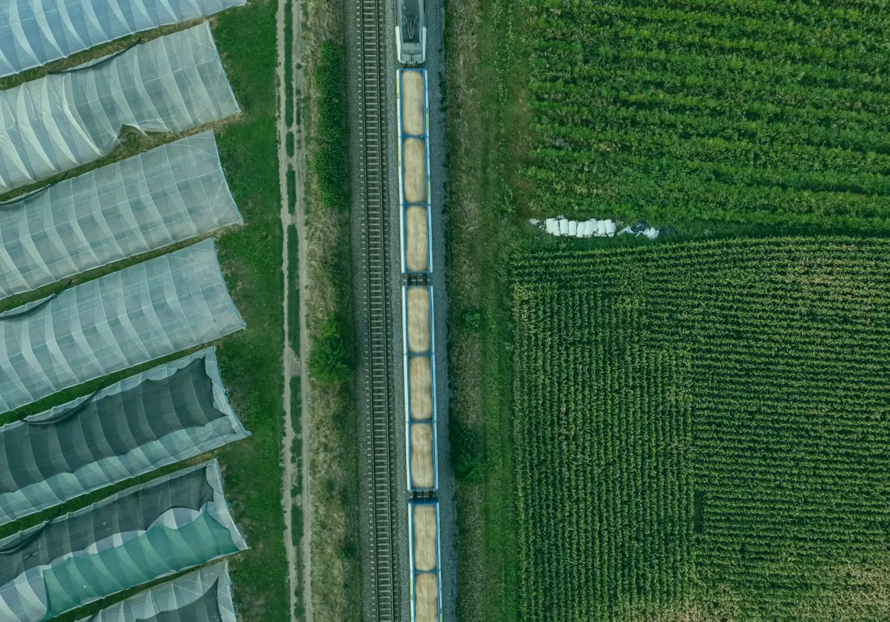 Aerial view of a freight train carrying grain between green farmland and covered crop structures, illustrating sustainable transport in agriculture.