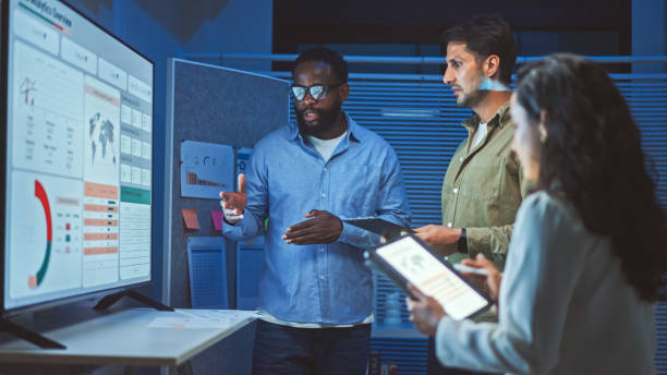 Three coworkers discuss data displayed on a large screen in an office at night.