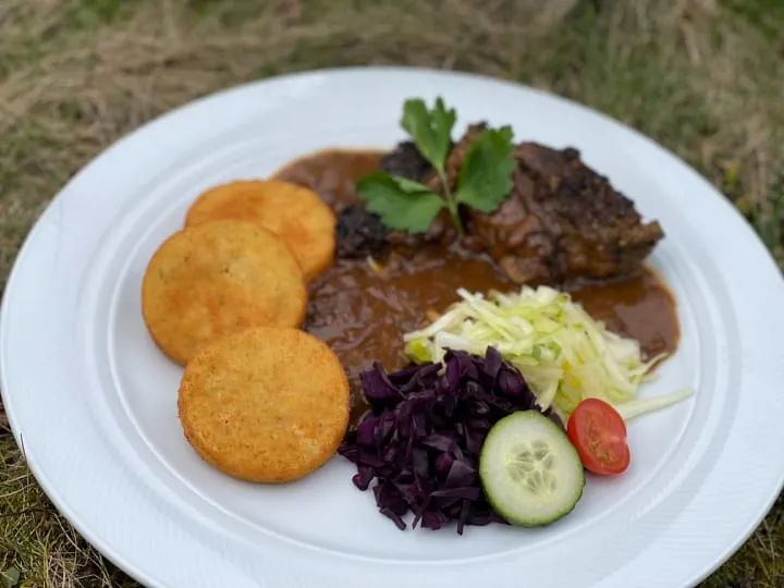 Mutzbraten mit Kroketten, Rotkohl und Gurkengarnitur auf weißem Teller.