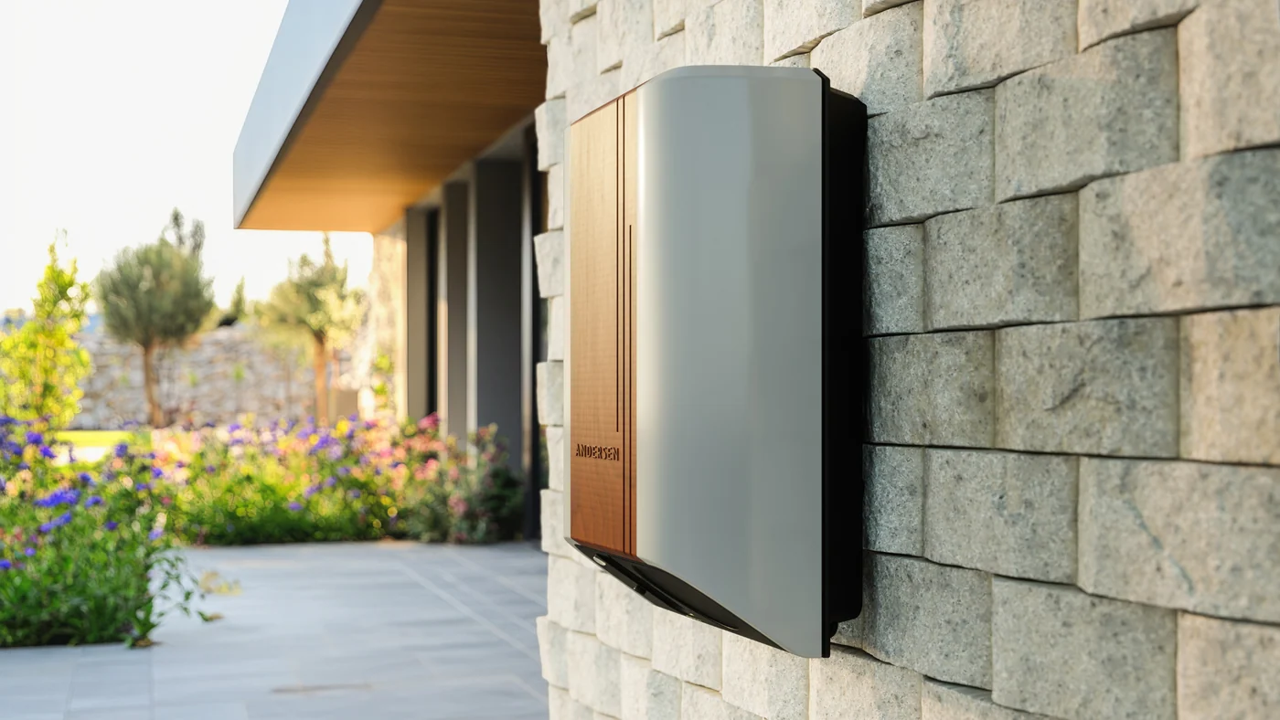 A modern Andersen electric vehicle charger with a grey casing and a wood-finish front panel, mounted on a textured, light-colored stone wall outside a house. A paved patio and a garden with purple flowers are visible in the soft-lit background