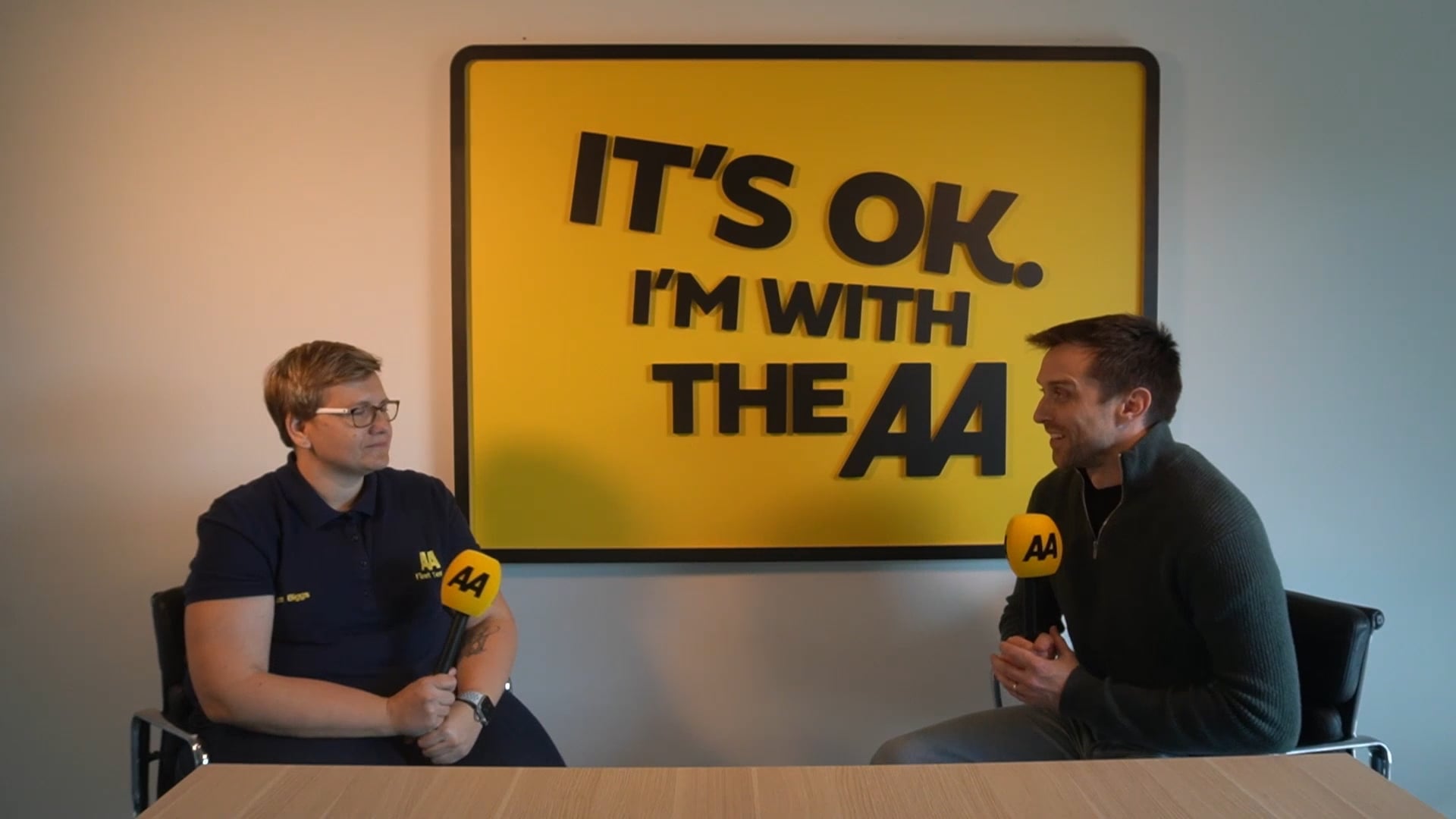 Sam Biggs, Fleet Manager at the AA, and Charlie, Rightcharge CEO, sitting at a desk during a video interview. Both are holding yellow AA-branded microphones, with a large yellow sign on the wall behind them reading 'IT'S OK. I'M WITH THE AA'.