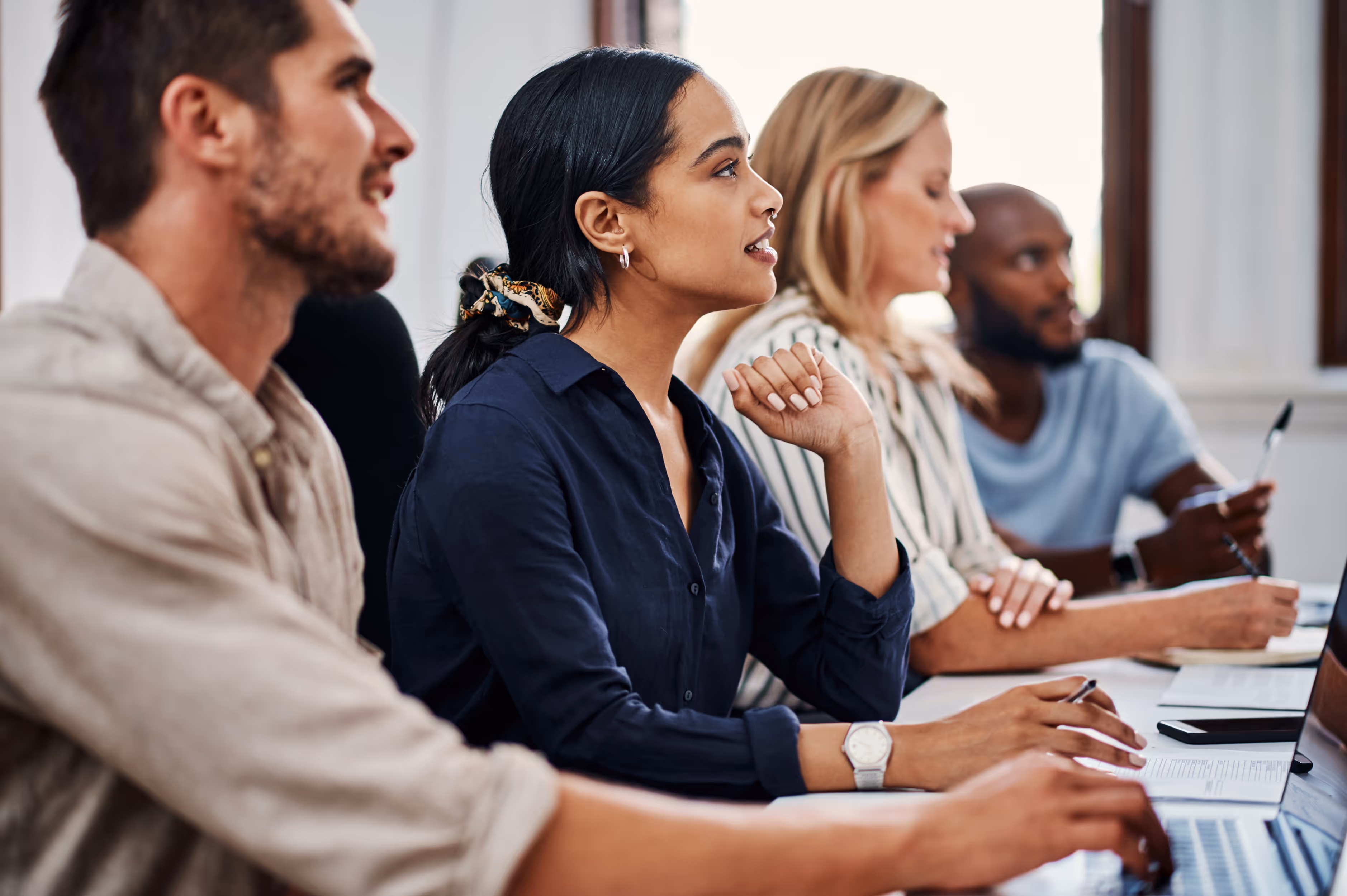 Four diverse young adults attentively participating in a meeting or class, seated side by side with laptops and notebooks.