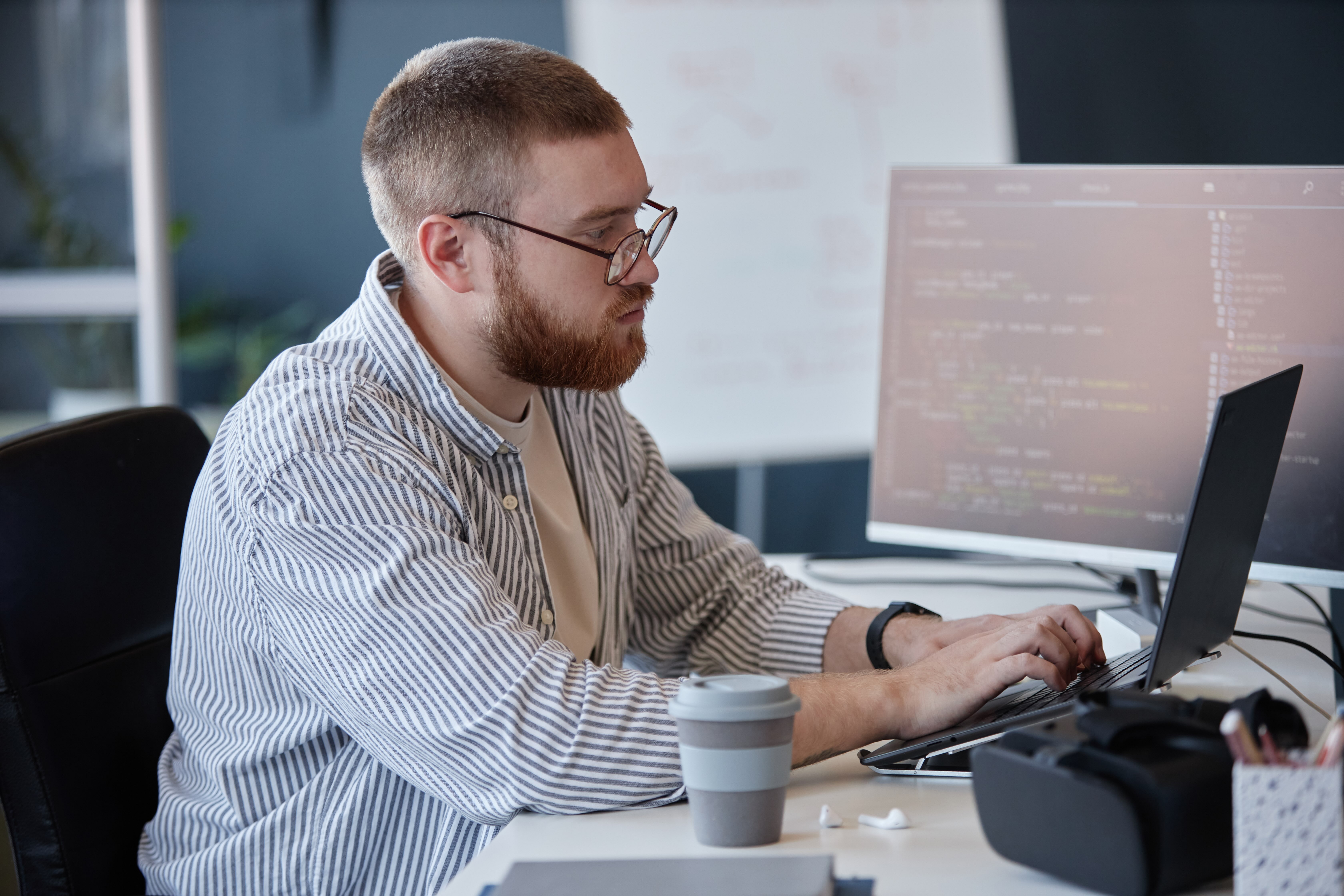 Man with glasses and beard typing on a laptop at a desk with a coffee cup and VR headset.