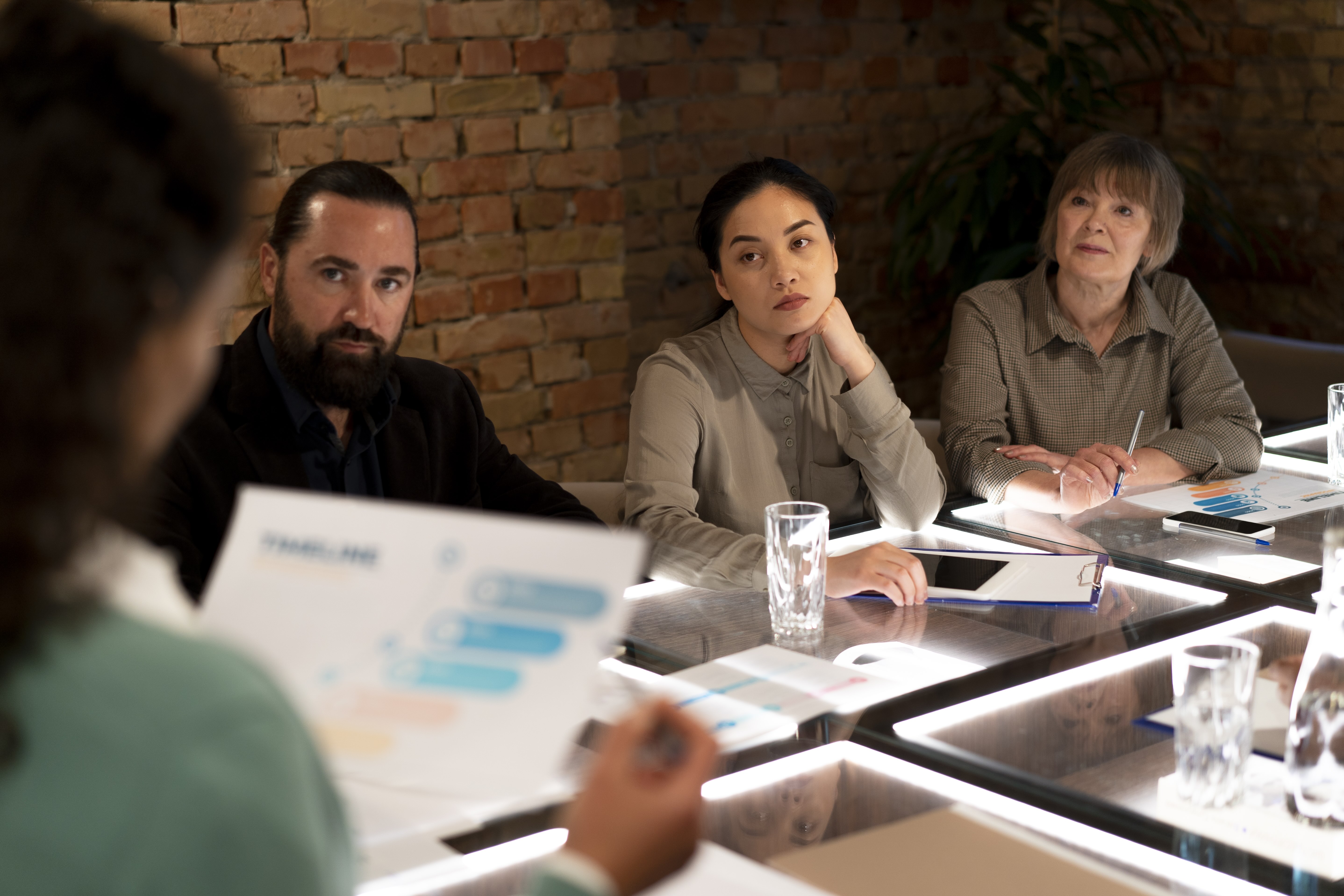 Three people attentively listening to a presenter holding a chart in a meeting room with exposed brick walls.