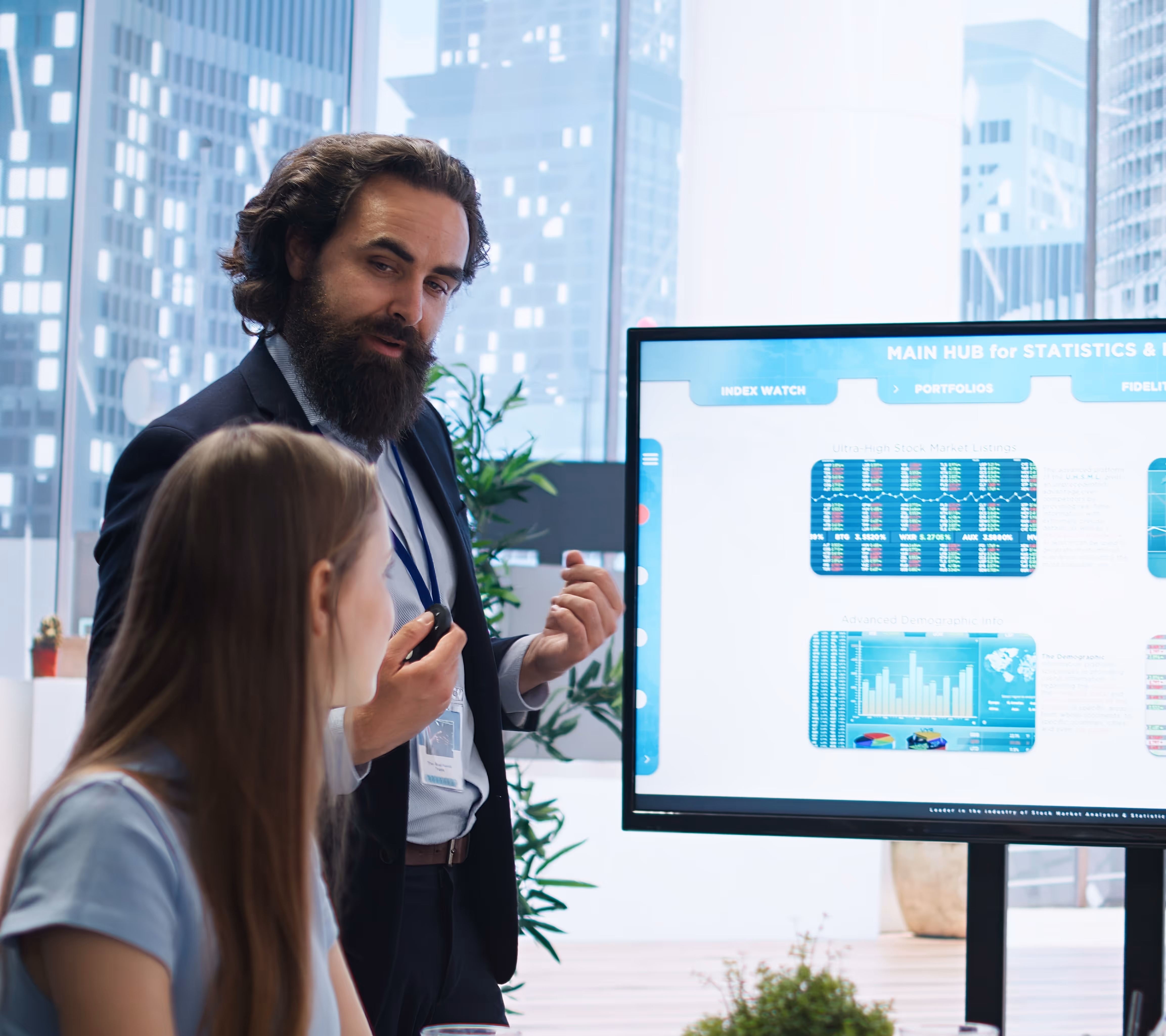 Bearded man in a suit explaining stock market data on a screen to a woman in a modern office.