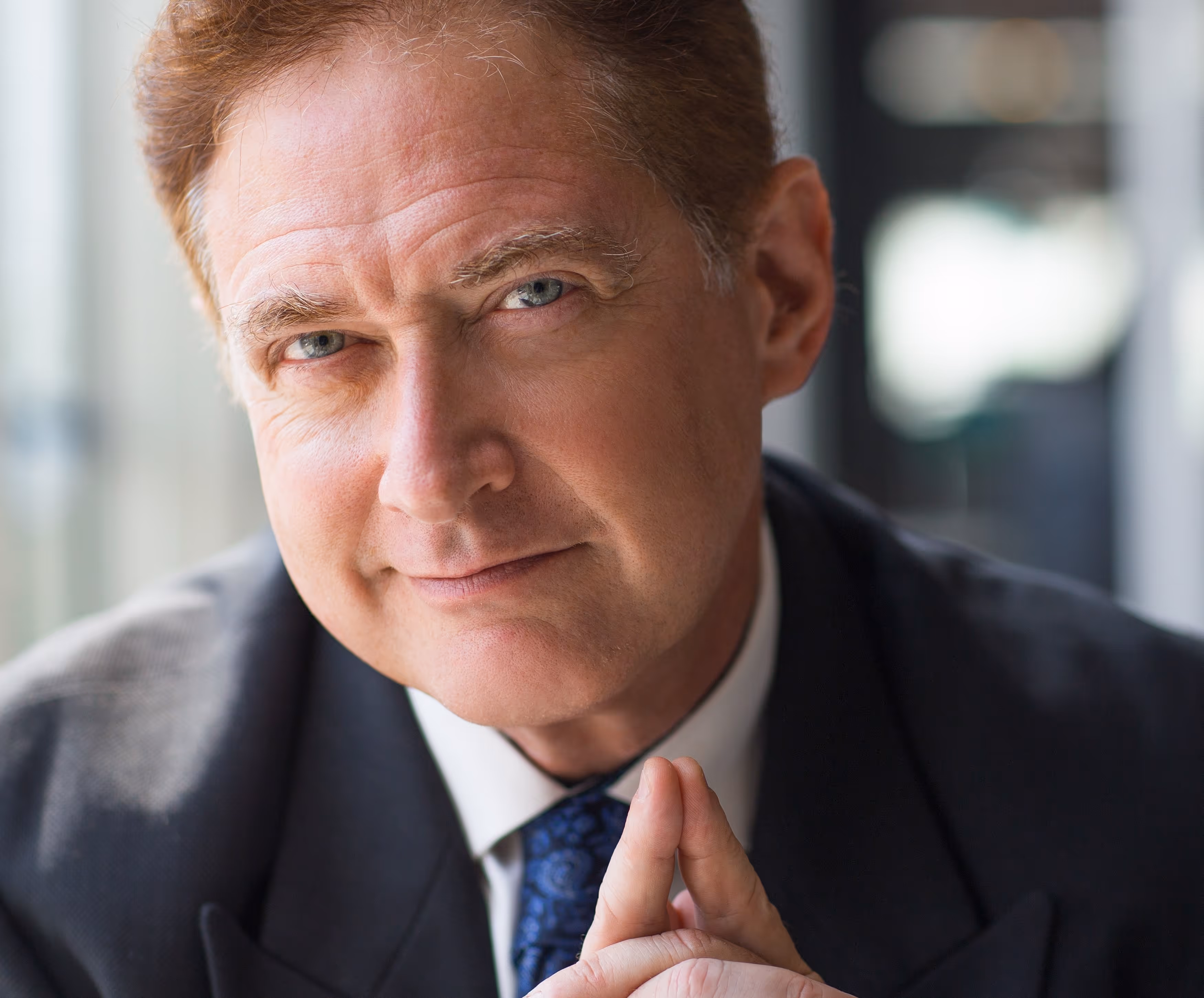 Close-up portrait of a confident middle-aged man in a suit with blue eyes and fingers touching in front of his face.