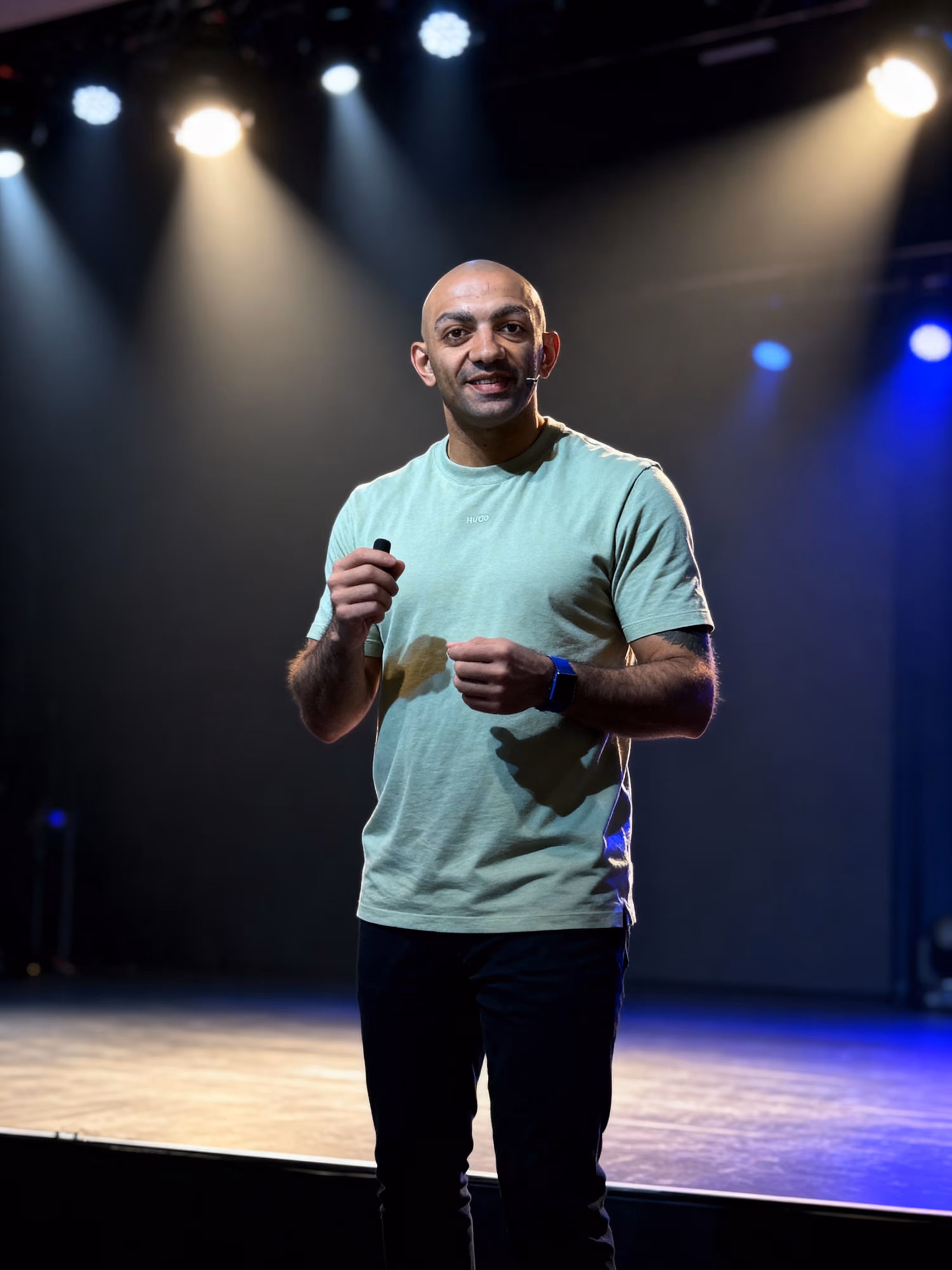 Bald man wearing a green Hugo t-shirt and black pants speaking on stage with a headset microphone and spotlight overhead.