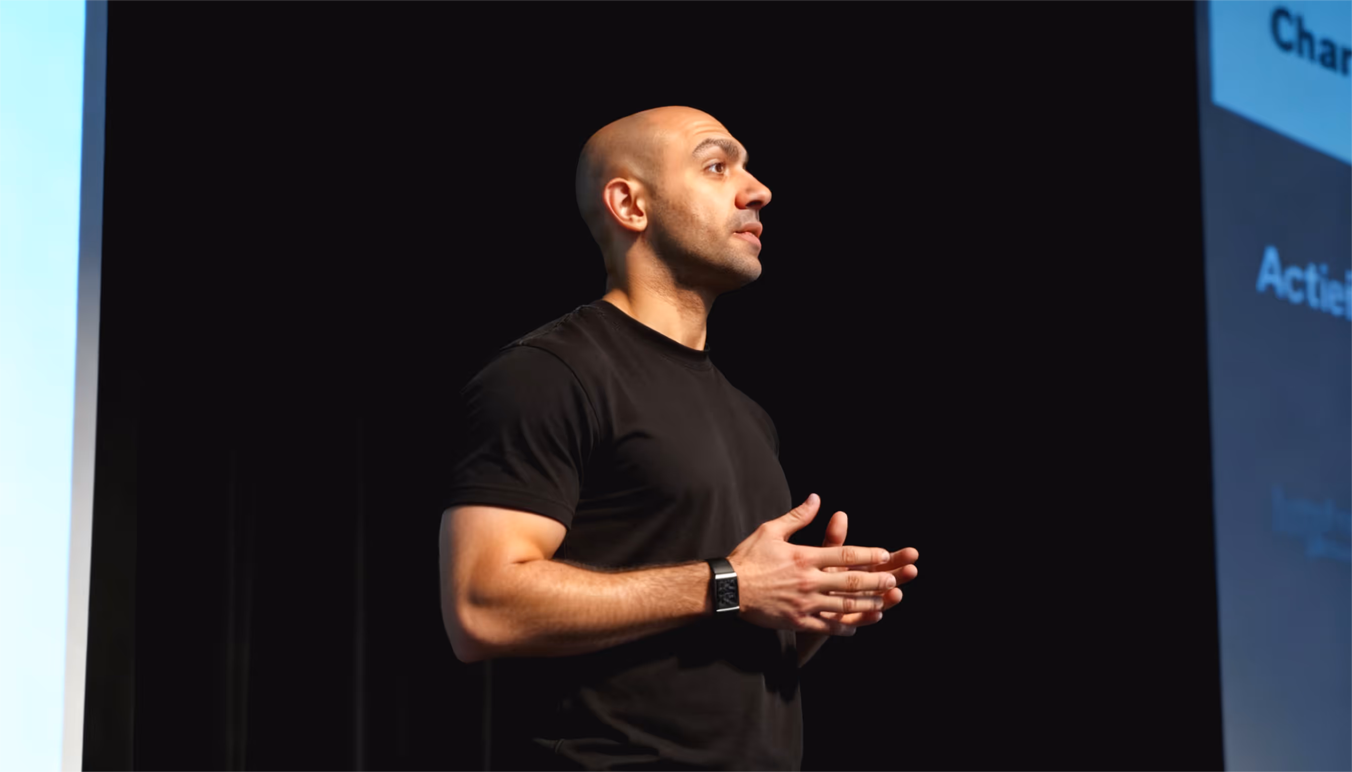 Yar in black t-shirt speaking on stage with dark background and presentation screen.