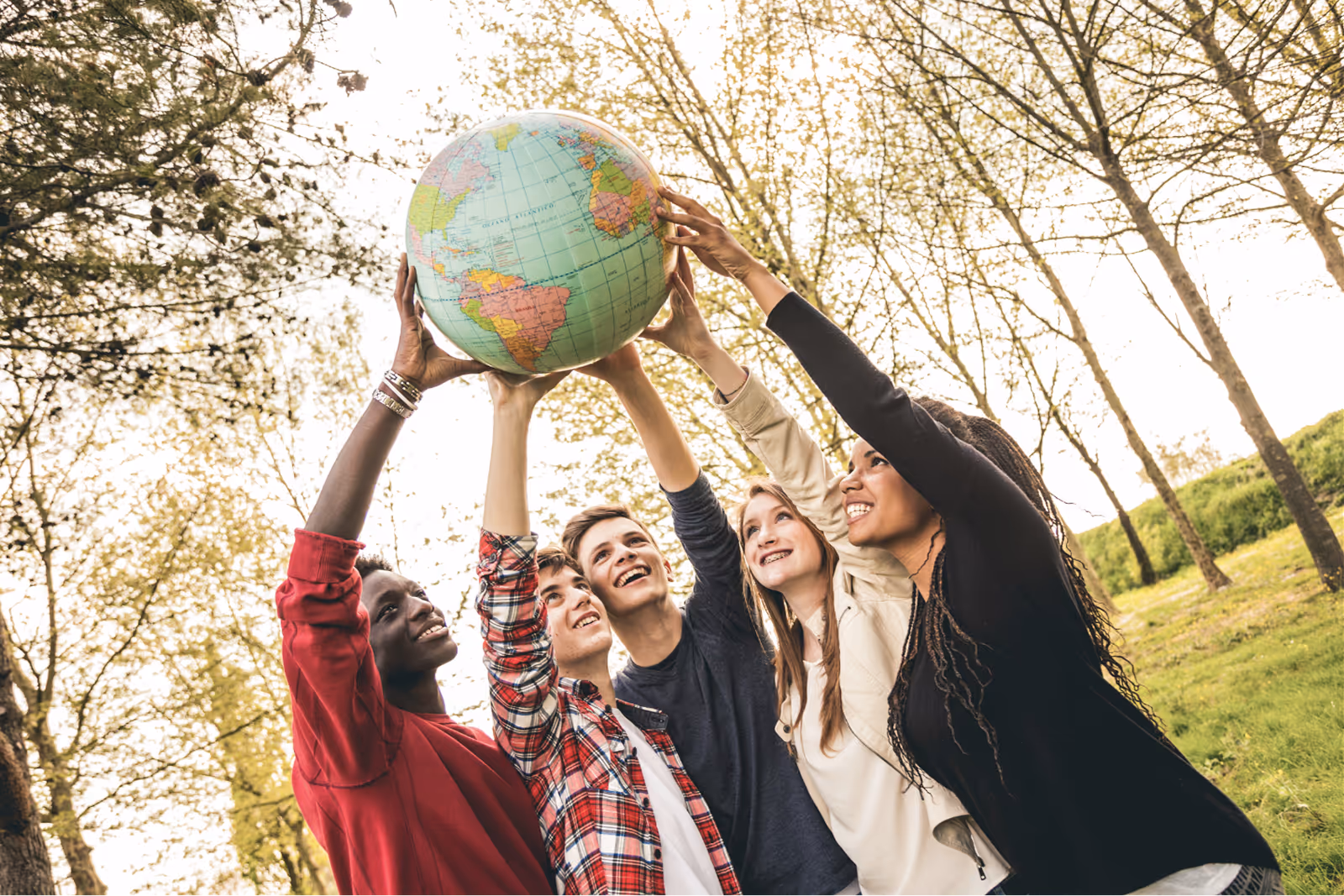 Four young people holding a globe above their heads together in a park.