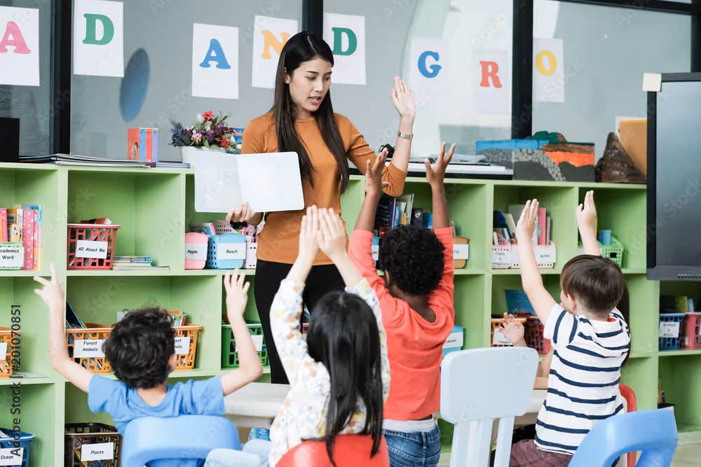 A teacher leading young children in a classroom activity with raised hands.