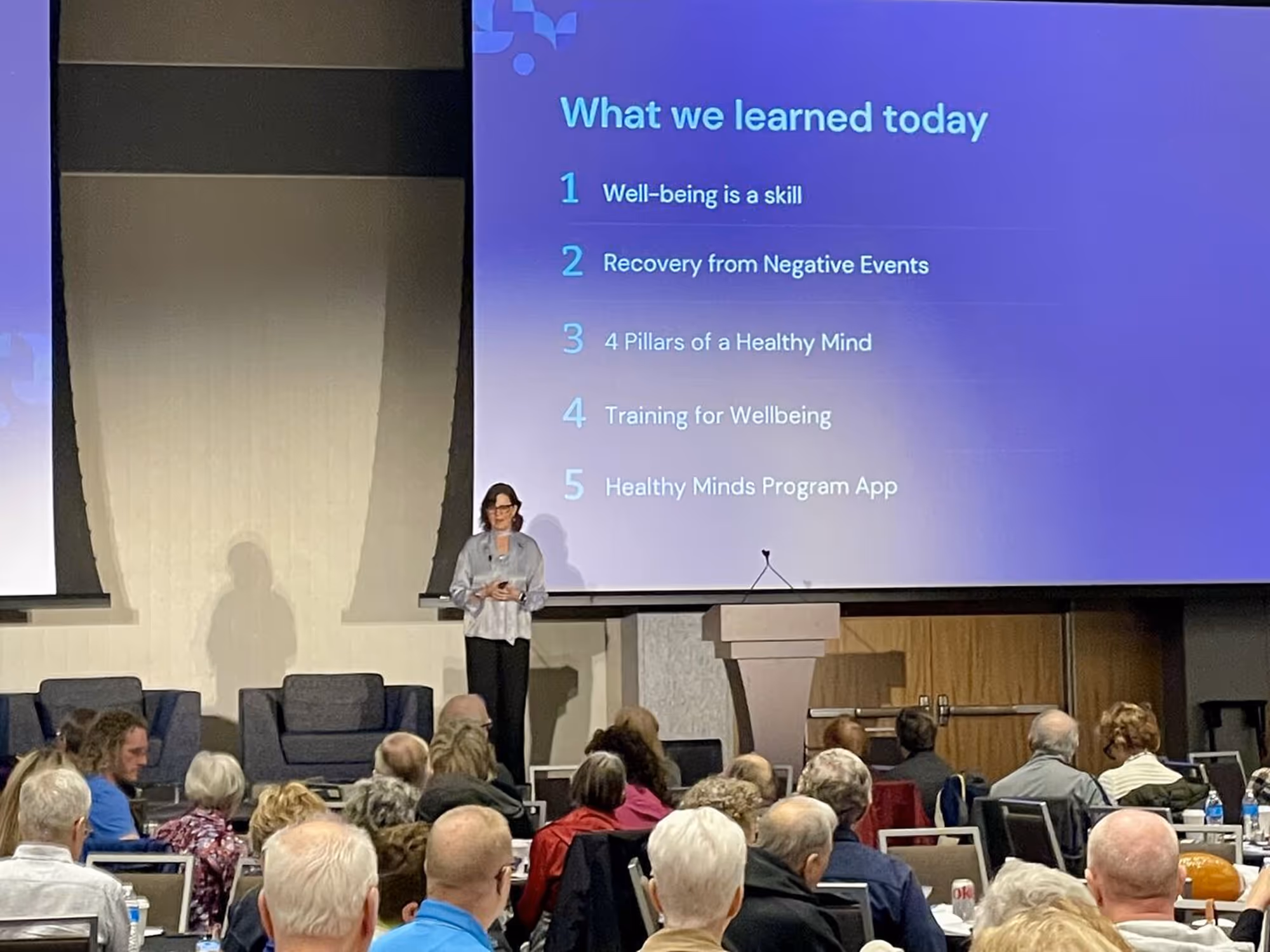 A speaker presenting to a large audience in a conference room with a slide behind her.