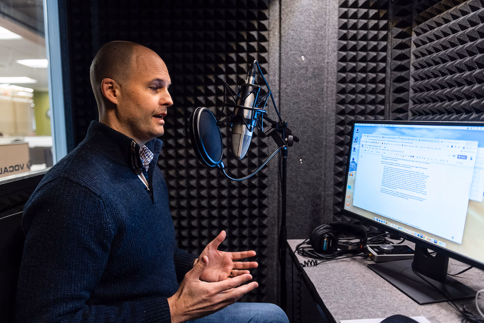 A man in a recording booth speaking into a microphone while reading from a screen.