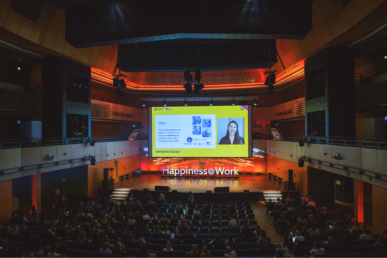 A large audience sits in a concert hall watching a presentation on a big screen during the Happiness@Work event.