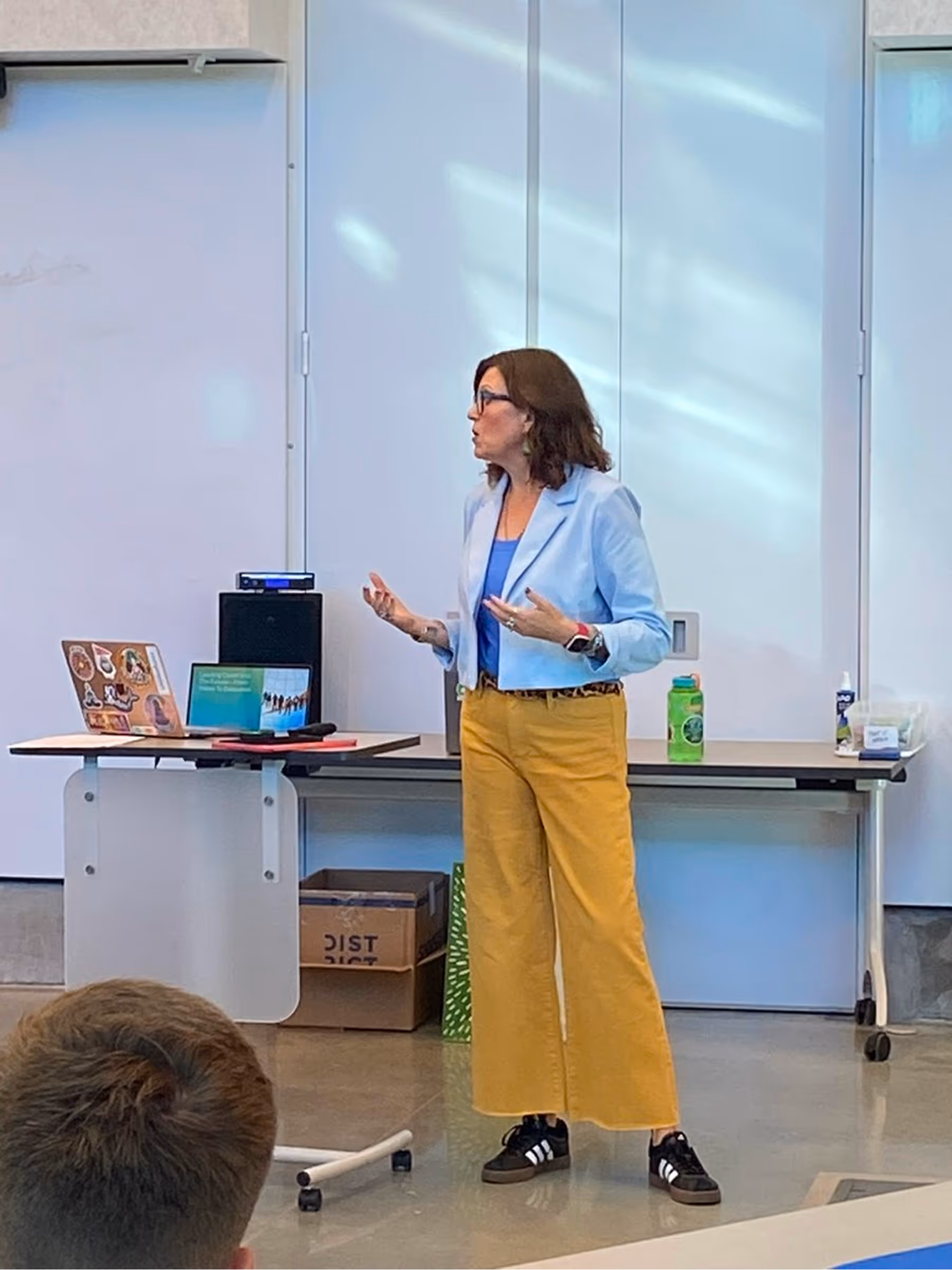 A woman speaking in front of a classroom during a presentation with a laptop on the desk.