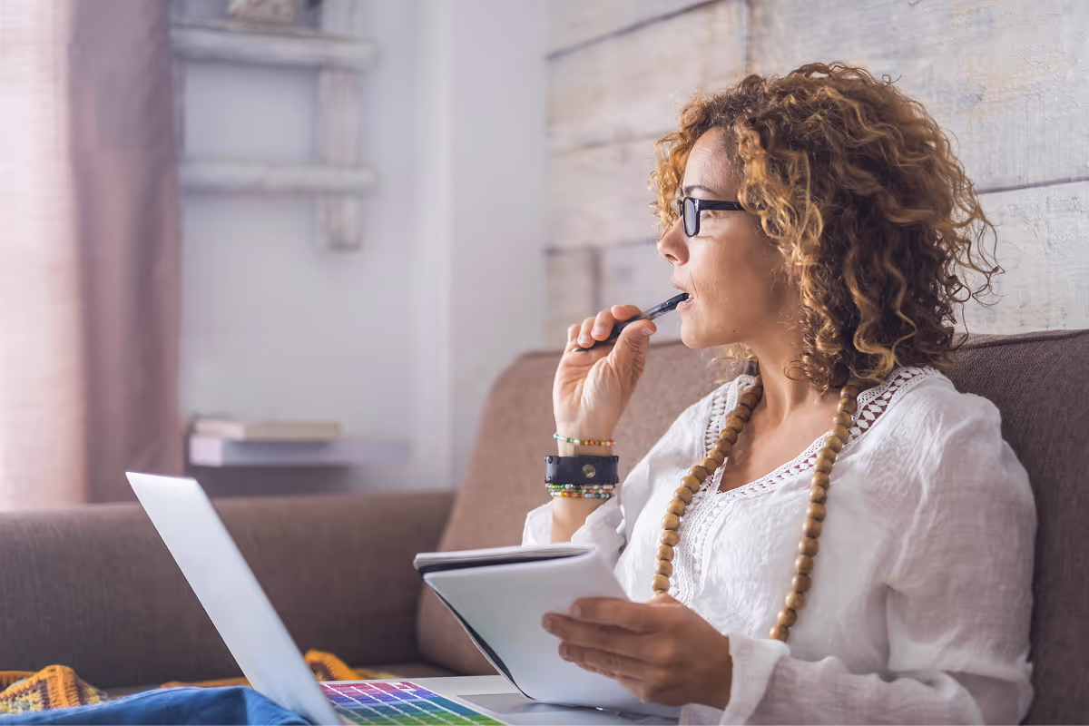 A woman sitting on a couch thinking while working on her laptop and notebook.