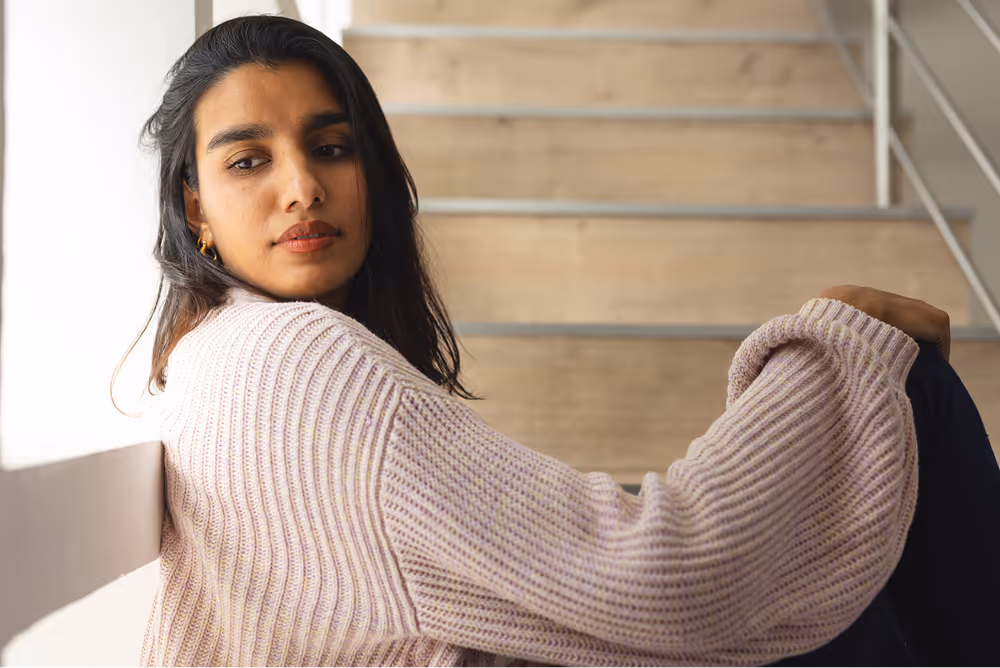 A woman sitting on wooden stairs looking out thoughtfully with soft natural light.
