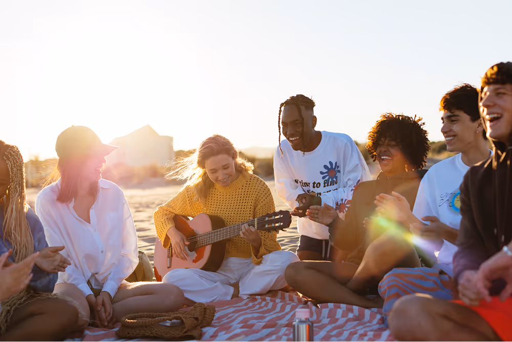 A group of friends sitting on the beach at sunset playing guitar and singing together.