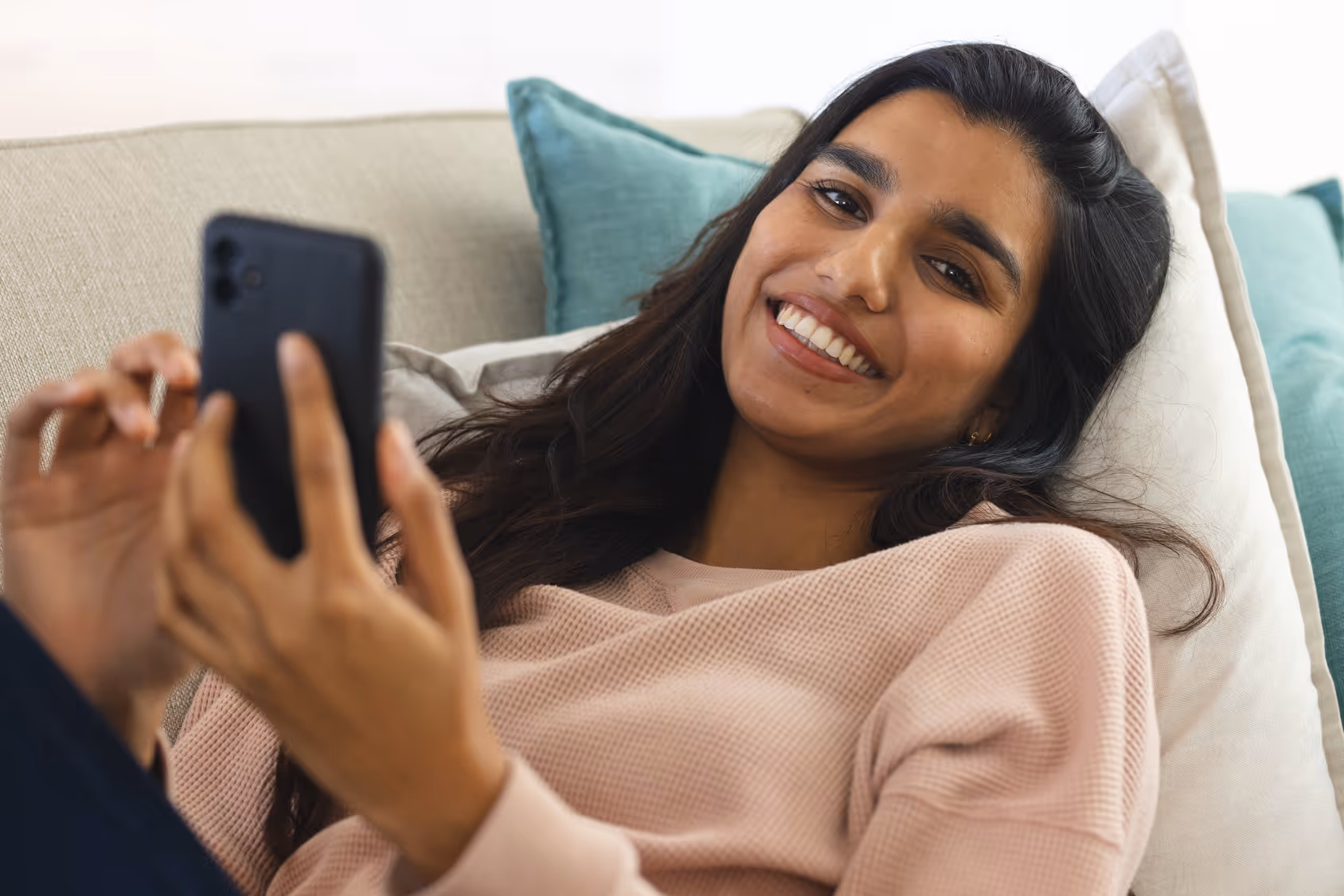 Smiling woman with long dark hair reclining on a couch using a smartphone.