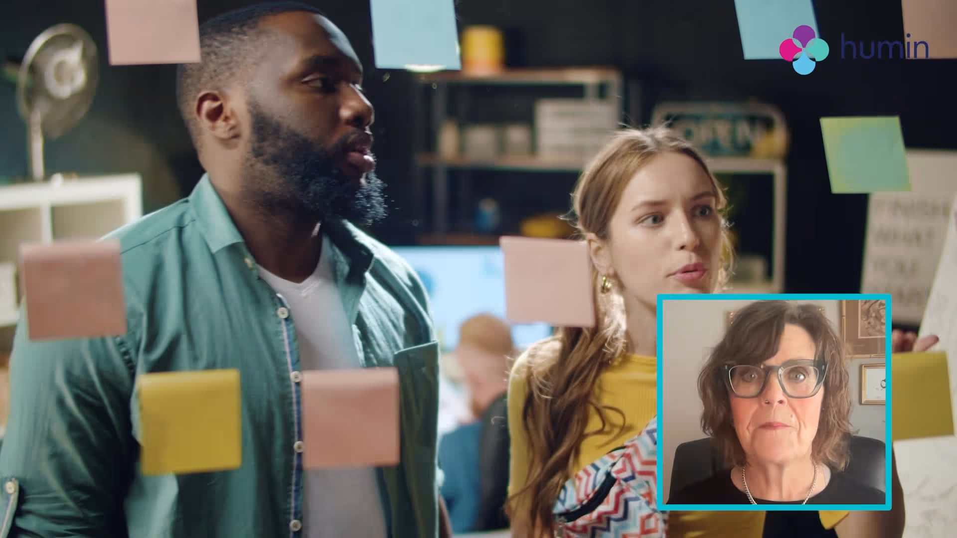 Two people discussing and pointing at sticky notes on a glass wall with an inset video call window showing a woman with glasses speaking.