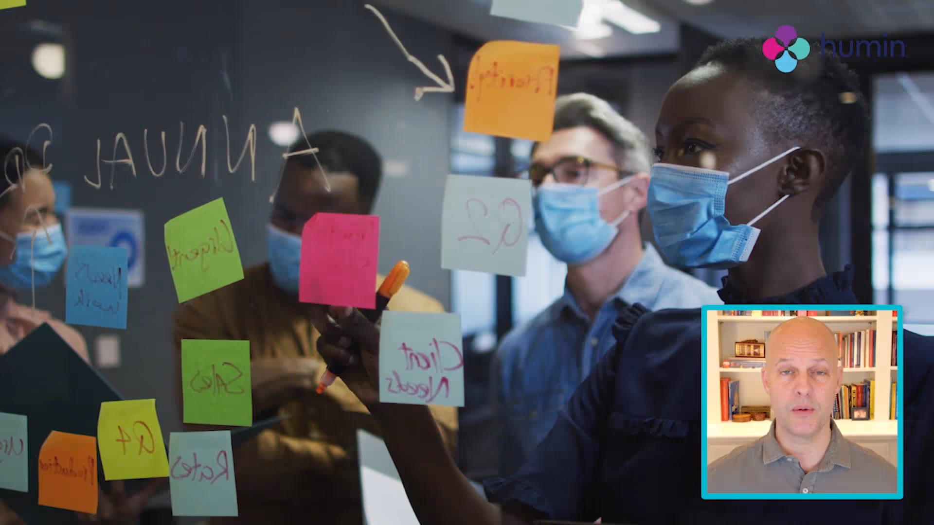 Team of masked colleagues brainstorming with colorful sticky notes on glass wall during a virtual meeting with a man shown in an inset video call window.