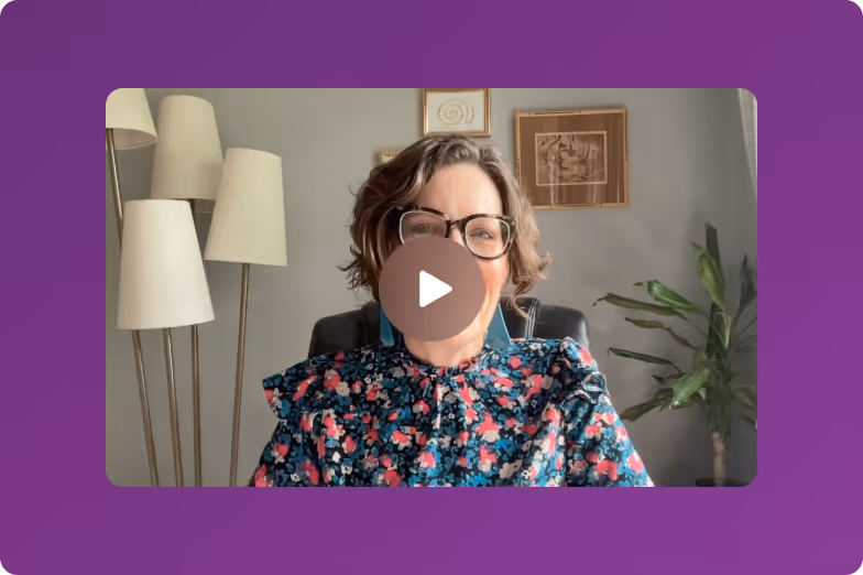 Woman with short hair and glasses smiling, wearing a floral patterned top, seated in a room with floor lamps and framed artwork on the wall.