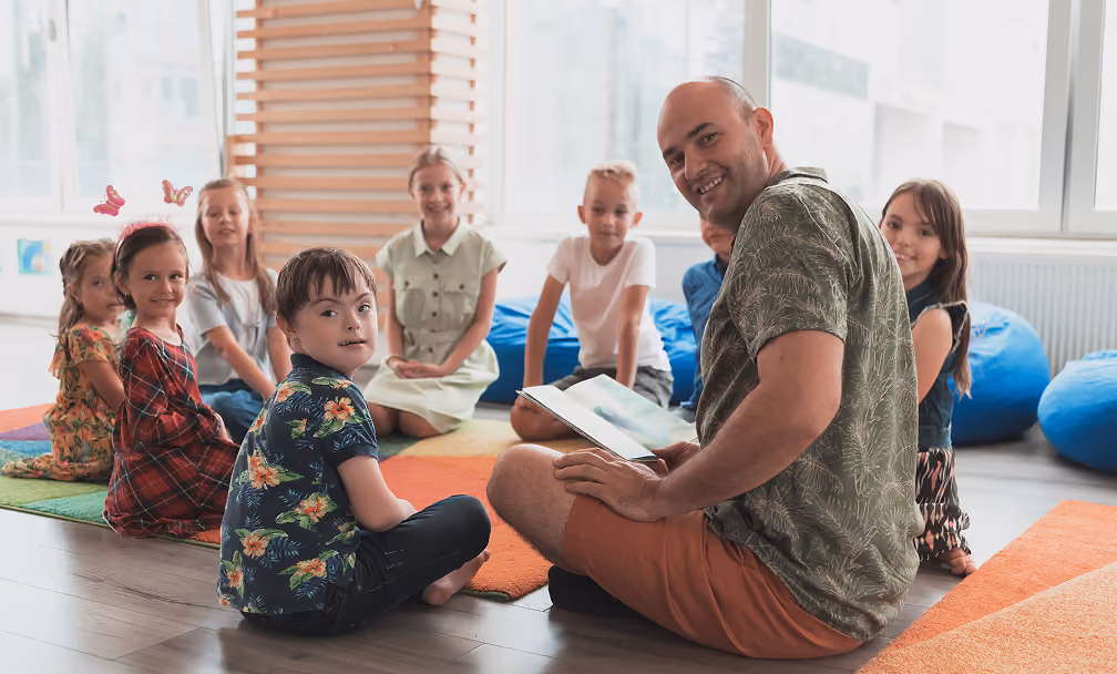 A teacher sitting on the floor reading a book to a group of children who are seated around him on colorful mats in a bright classroom with large windows.