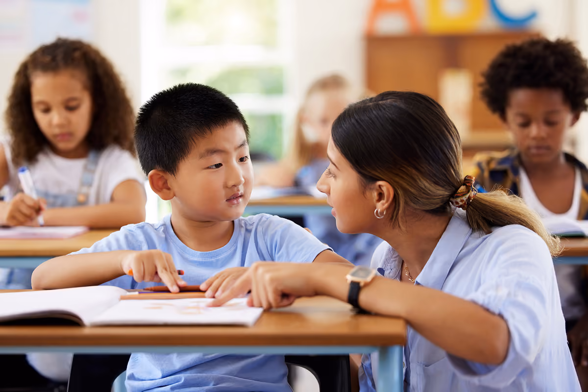 Teacher leaning over a student's desk, providing assistance while the student looks at her attentively in a classroom with other children working at their desks.