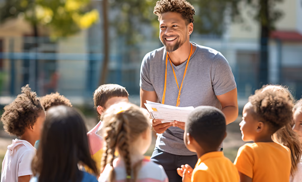 Smiling male teacher holding papers and engaging with a diverse group of young children outdoors with trees and a building in the background.