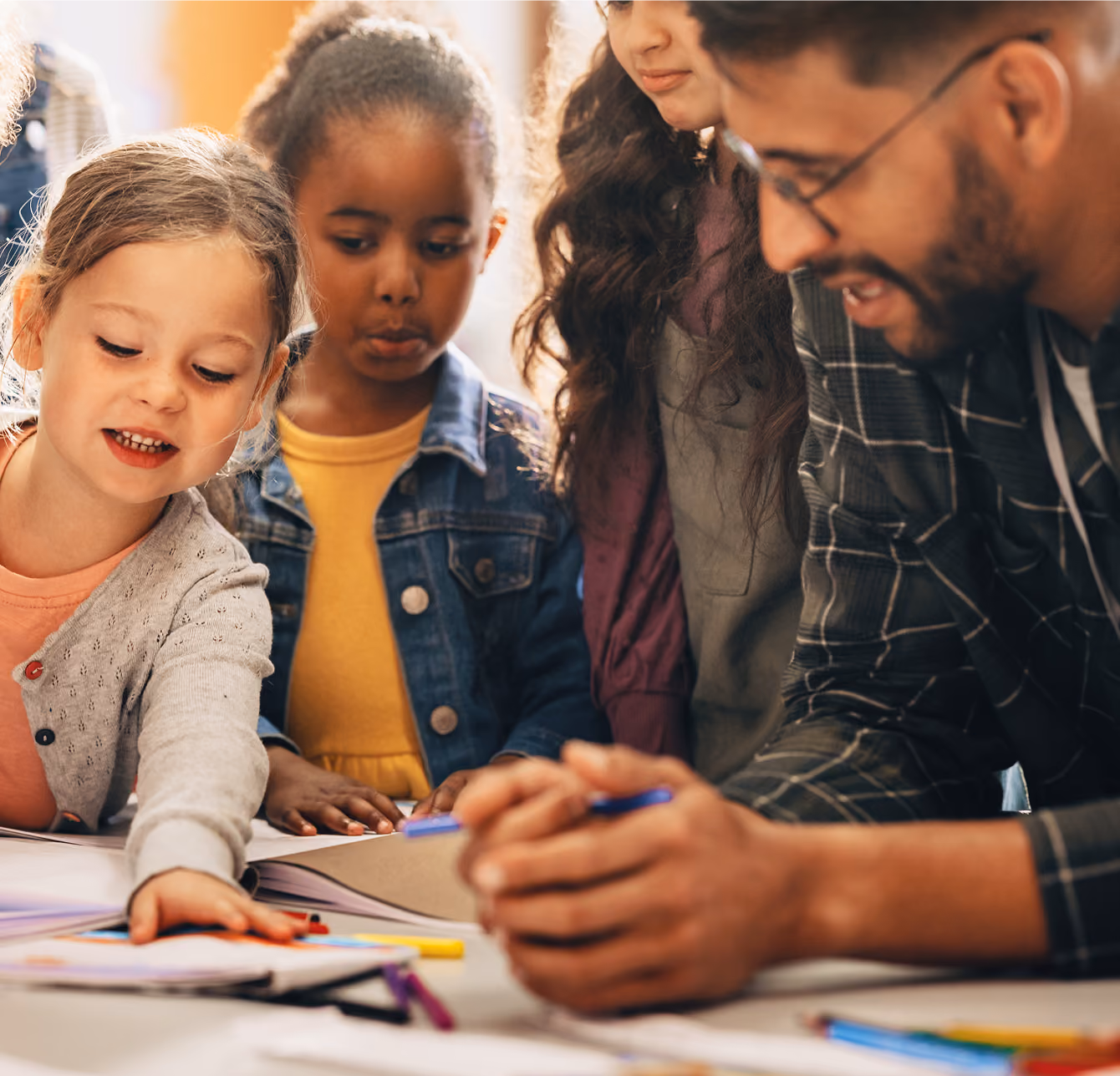 A teacher with glasses and a checkered shirt is leaning over a table, engaging with three young children focused on drawing and coloring activities.