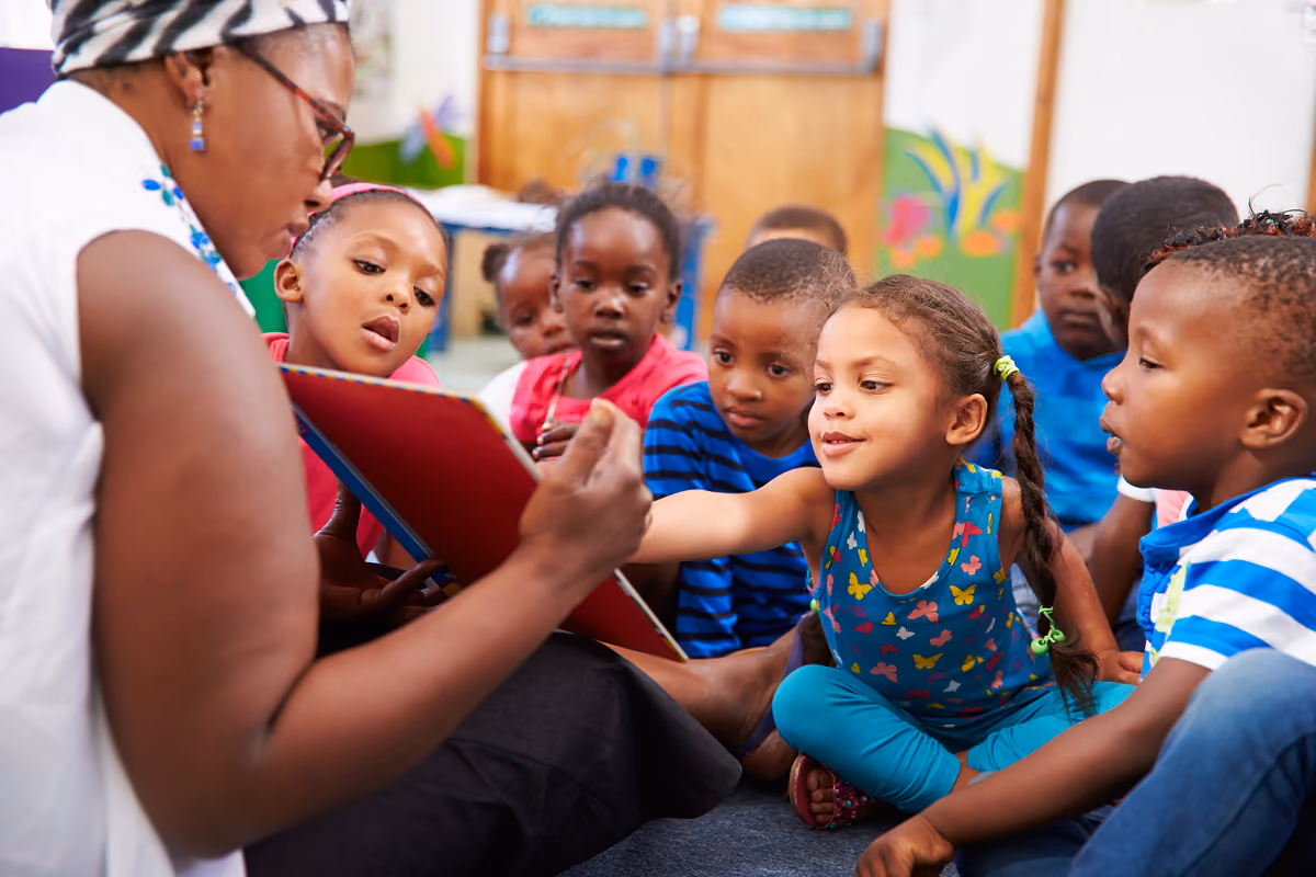 Teacher reading from a red book to a group of young children sitting on the floor, who are attentively looking and reaching towards the book in a classroom setting.