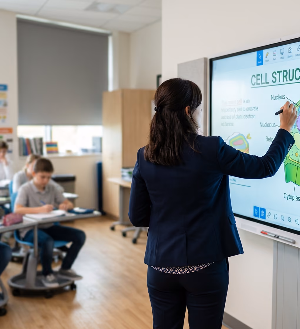 Teacher in a classroom writing on a large interactive screen displaying a diagram of cell structure, with students sitting at desks in the background.