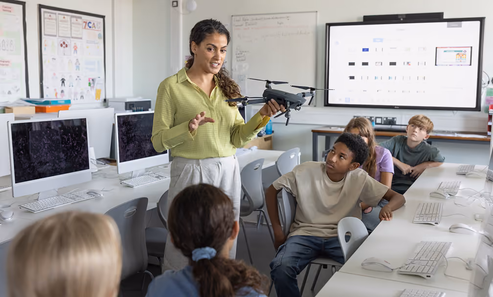 A female teacher holds a drone and explains its features to a group of students seated around computer desks in a classroom technology lab.