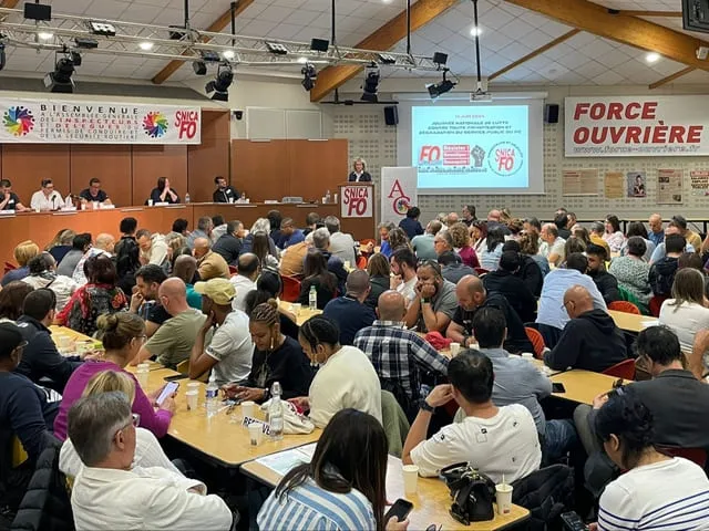 Une salle comble avec des personnes assises à des tables, assistant à une réunion ou assemblée avec un panneau SNICA FO et un écran de présentation à l'avant.