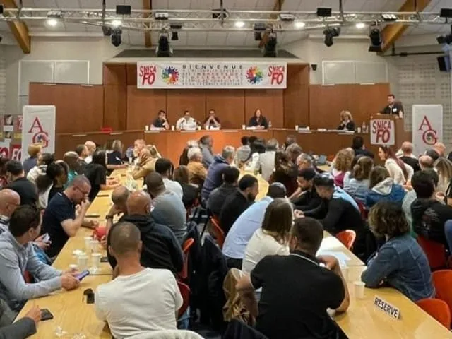 Grande assemblée générale avec des participants assis à des tables longues, un panel de cinq personnes sur une estrade et un orateur debout à un pupitre marqué SNICA FO.