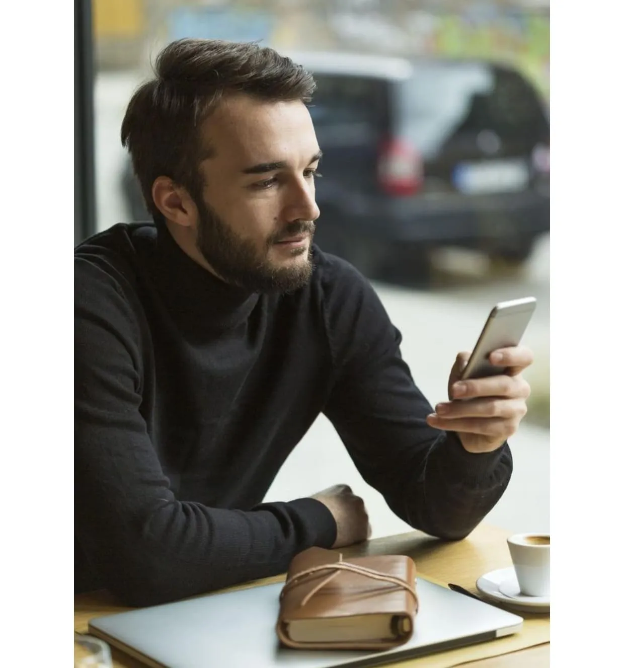 Un homme avec un pull noir regarde son téléphone assis à une table avec un carnet, un ordinateur portable fermé et une tasse de café.
