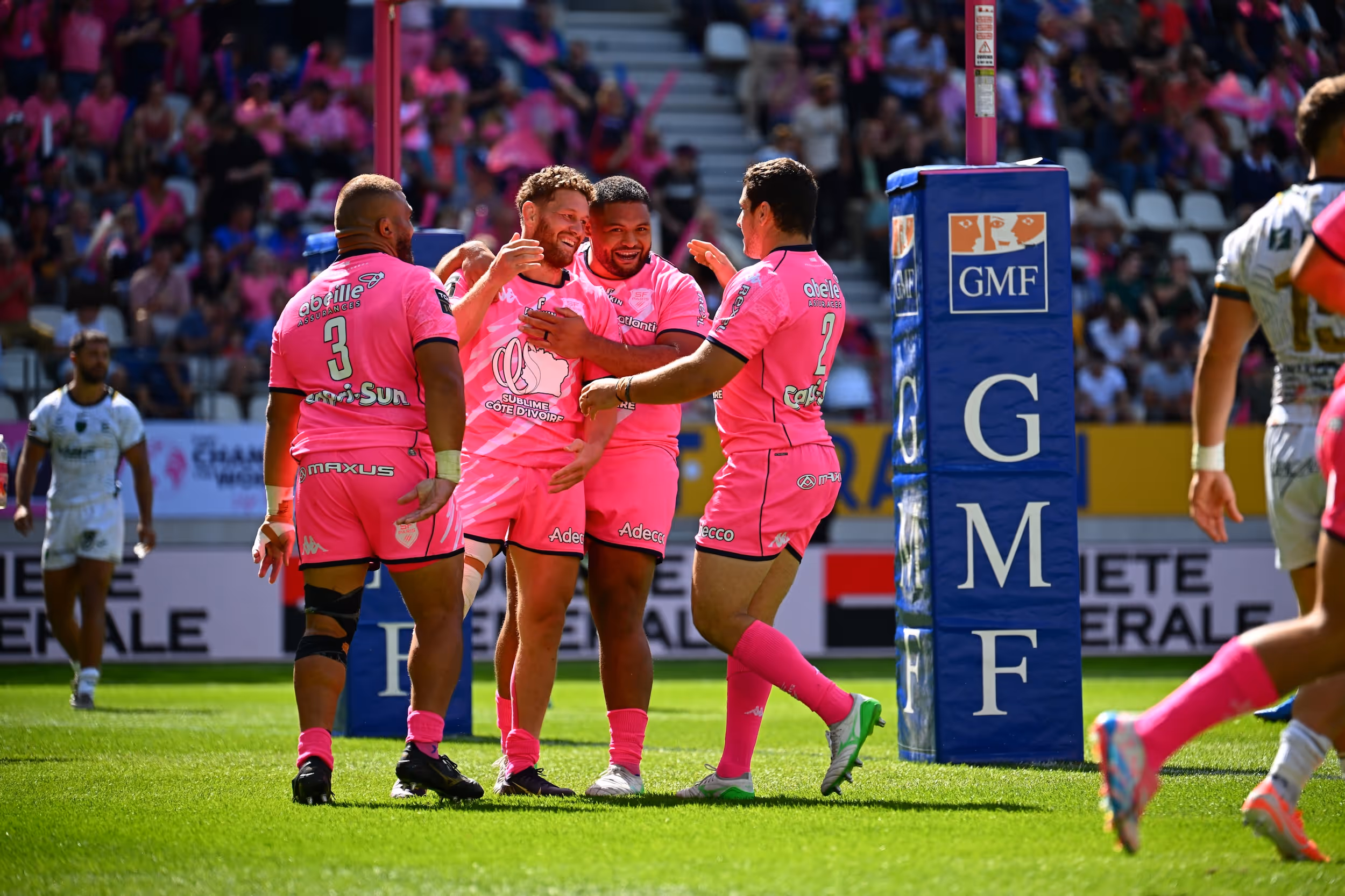 Rugby players in bright pink uniforms celebrating near the goal post on a sunny field with spectators in the background.