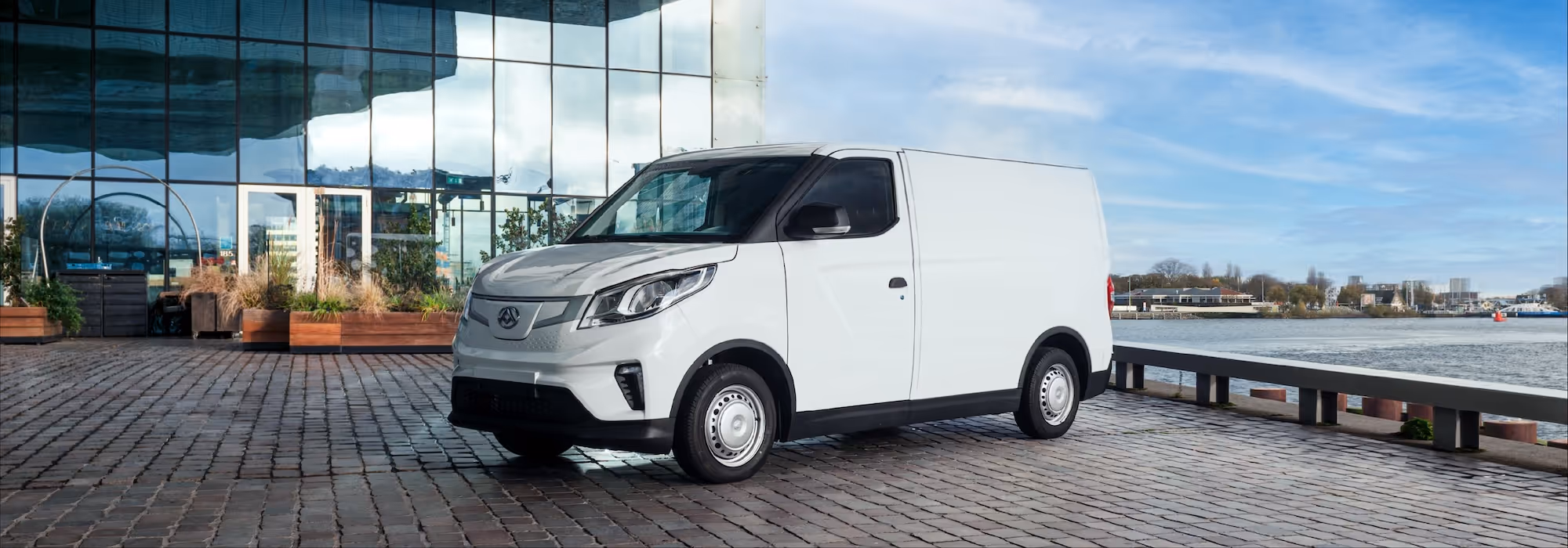 White cargo van parked on a cobblestone surface near a glass building and waterfront under a blue sky.