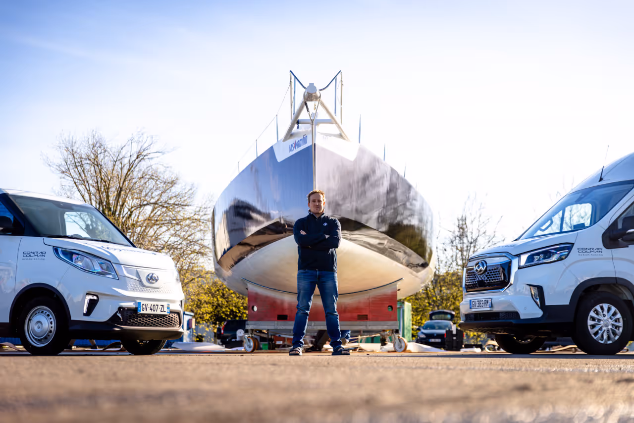 Man standing with arms crossed in front of a large boat hull on a trailer, flanked by two white vans with Conrad Colman Ocean Racing logos.
