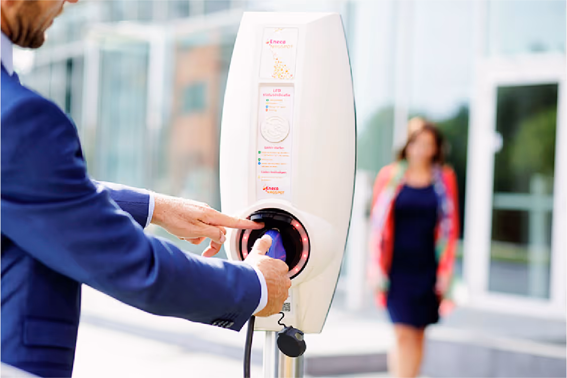 Man in suit plugging an electric vehicle charger into a charging station outdoors with a woman blurred in the background.